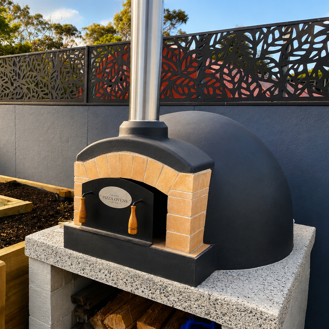 Outdoor pizza oven with a black dome and brick arch, mounted on a concrete countertop, with a stainless steel chimney and a decorative black metal fence in the background.
