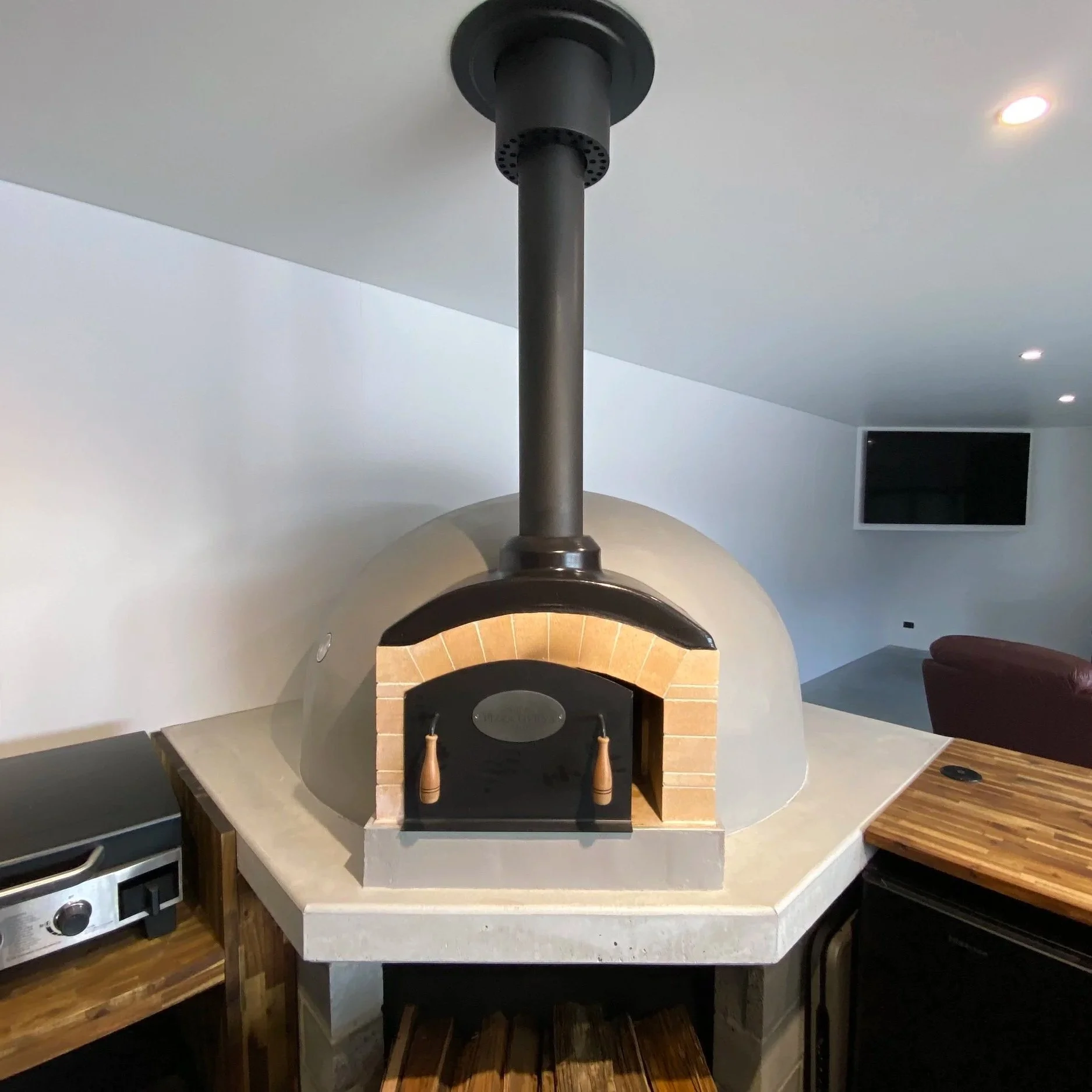 Indoor wood-fired pizza oven with brick arch and chimney pipe, situated on a white countertop in a modern kitchen.