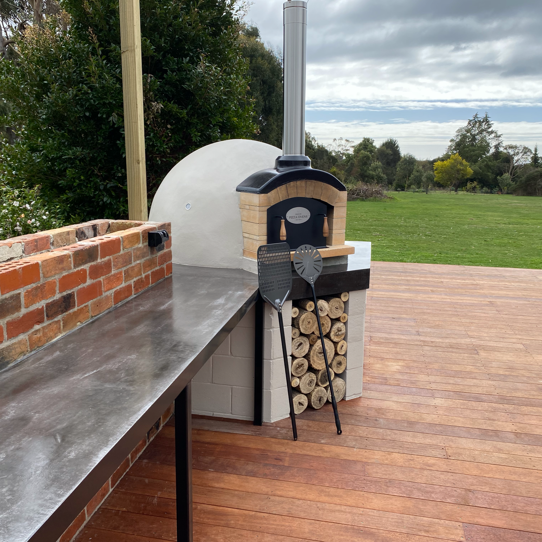 An outdoor pizza oven with a pizza oven sign, placed on a brick and metal countertop, with two pizza peel tools leaning against it, storage logs underneath, and a backdrop of green grass, trees, and a cloudy sky.