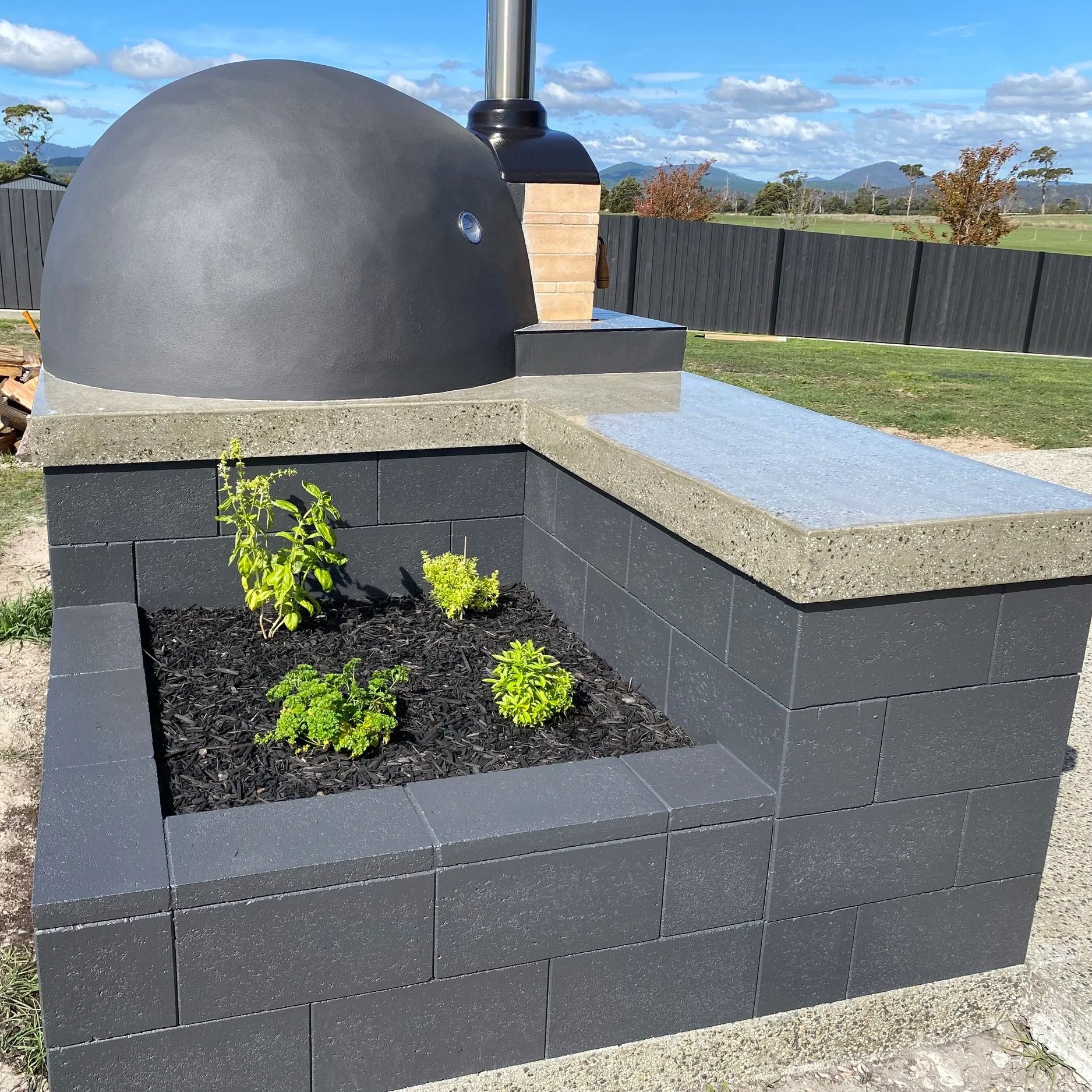 A modern outdoor pizza oven made of dark gray bricks with a scenic mountain view in the background.