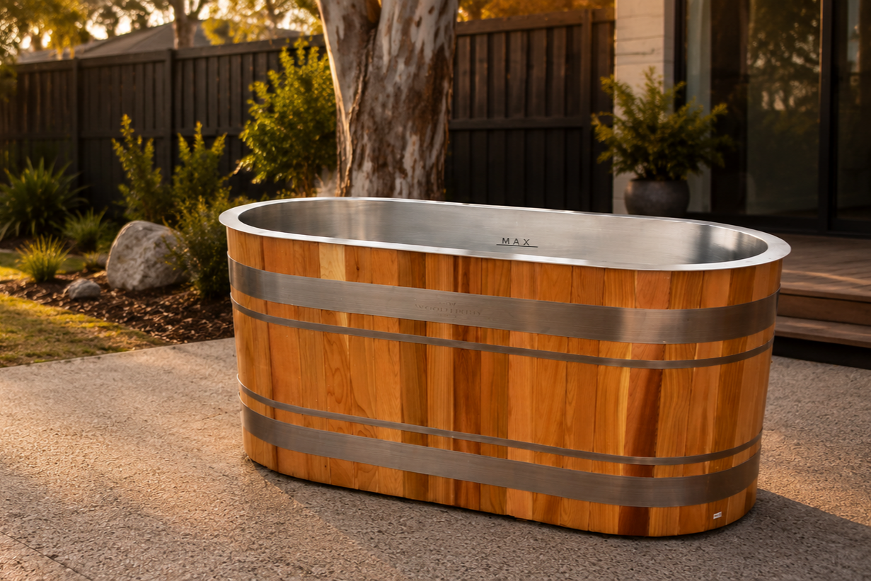 A wooden ice bath with stainless steel accents, placed outdoors on a patio with trees, plants, and a black fence in the background.