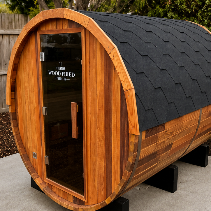 A wooden barrel-shaped sauna with a glass door and black shingle roof, placed on a concrete surface outside, with trees and a wooden fence in the background.