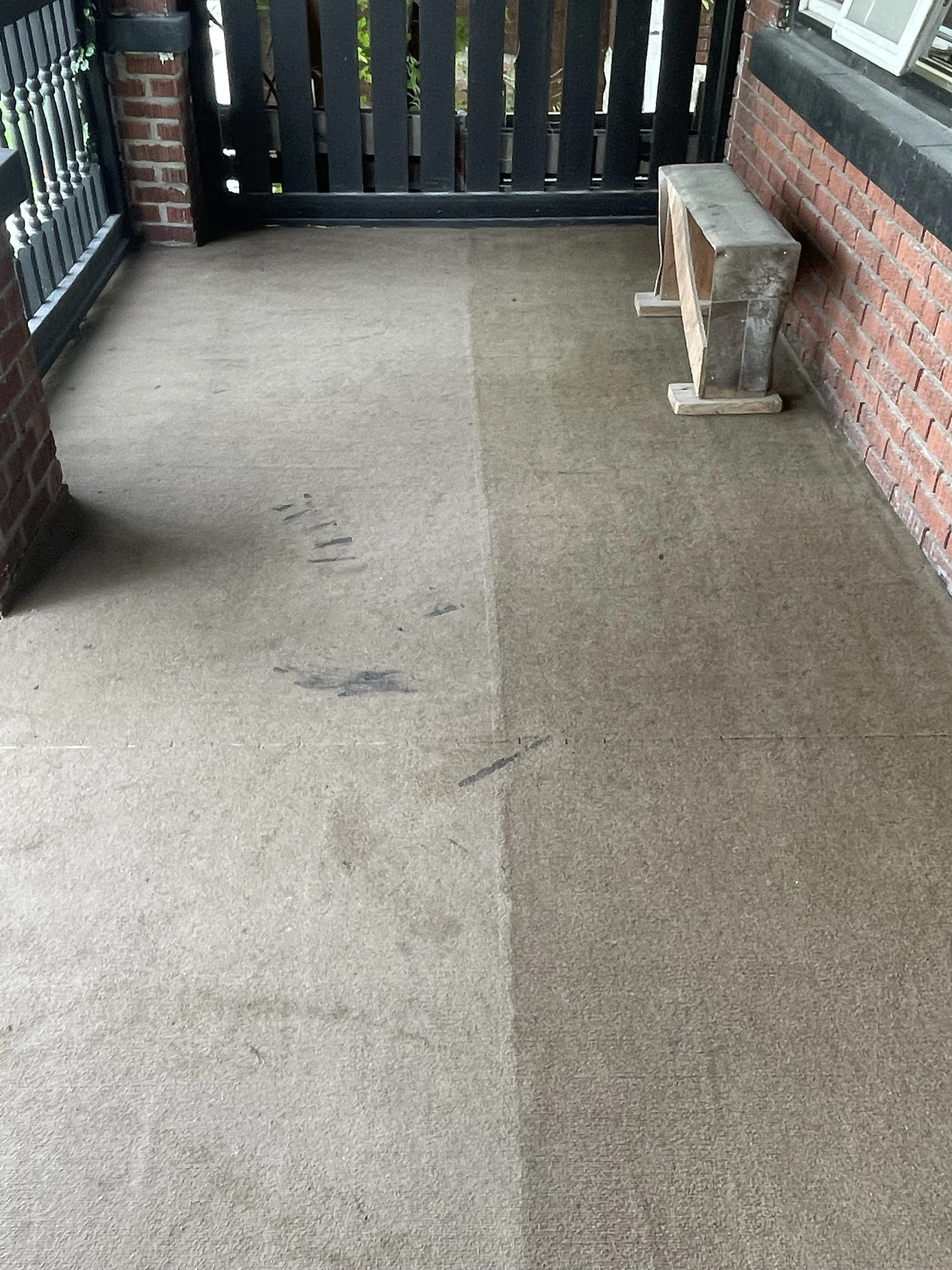 A small balcony with a concrete floor, brick walls, a black metal gate, and a small bench.