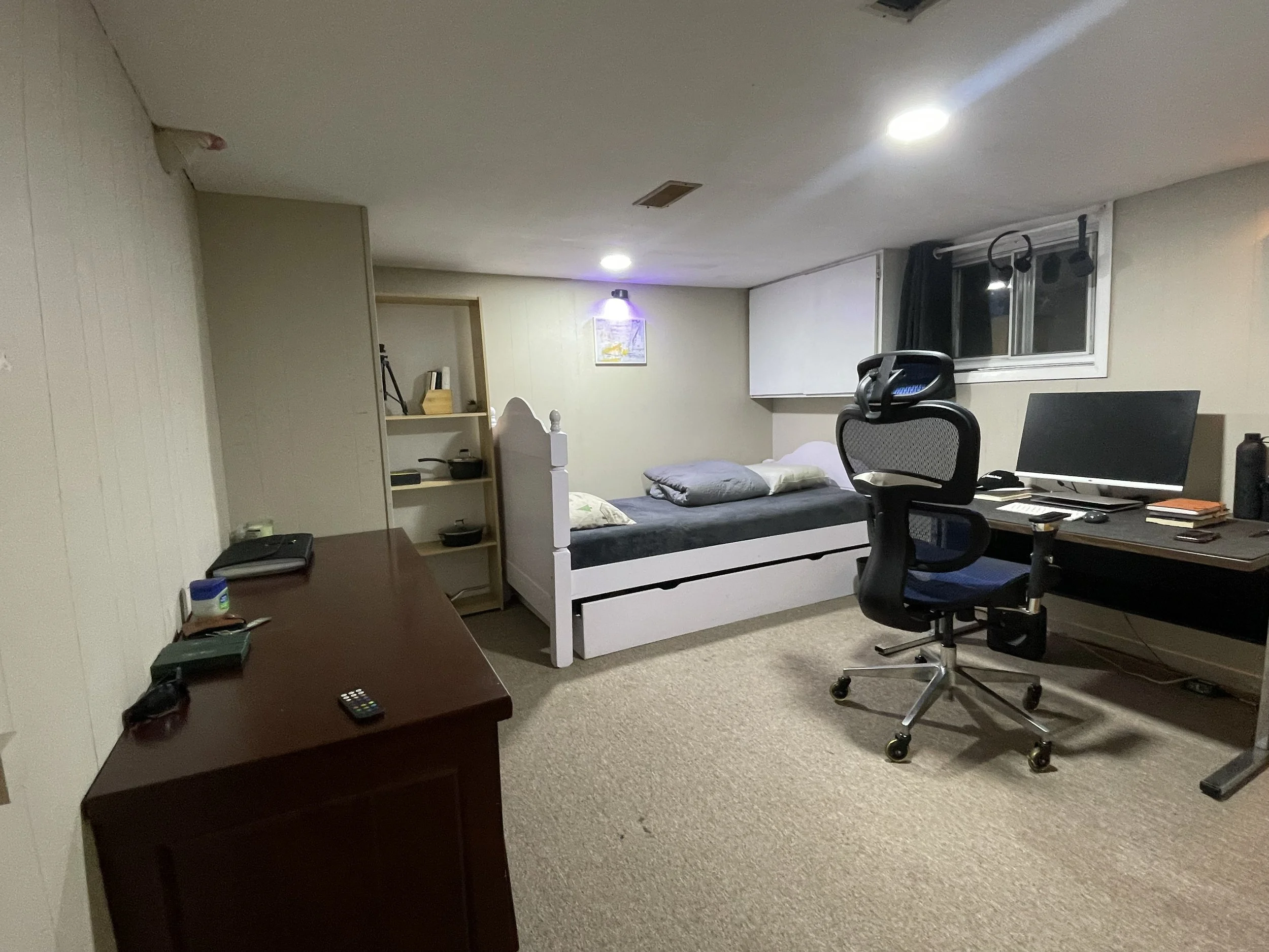 A room with beige carpet, a bed with pillows, a desk with a computer, a black office chair, a wooden dresser with various items, a bookshelf, and a window with black curtains.