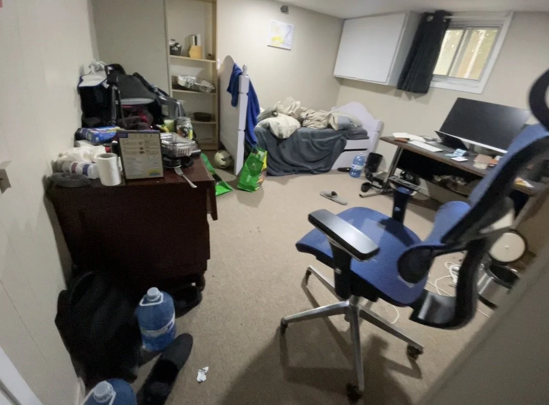 A cluttered office room with a desk, a blue office chair, a small white sofa with blankets, a computer monitor, and various items scattered around including cleaning supplies, a water bottle, and a backpack.