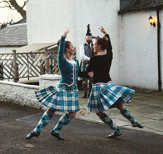 Two women in traditional Scottish Highland Dance costumes performing a propelled pivot turn with a bagpiper in the background.
