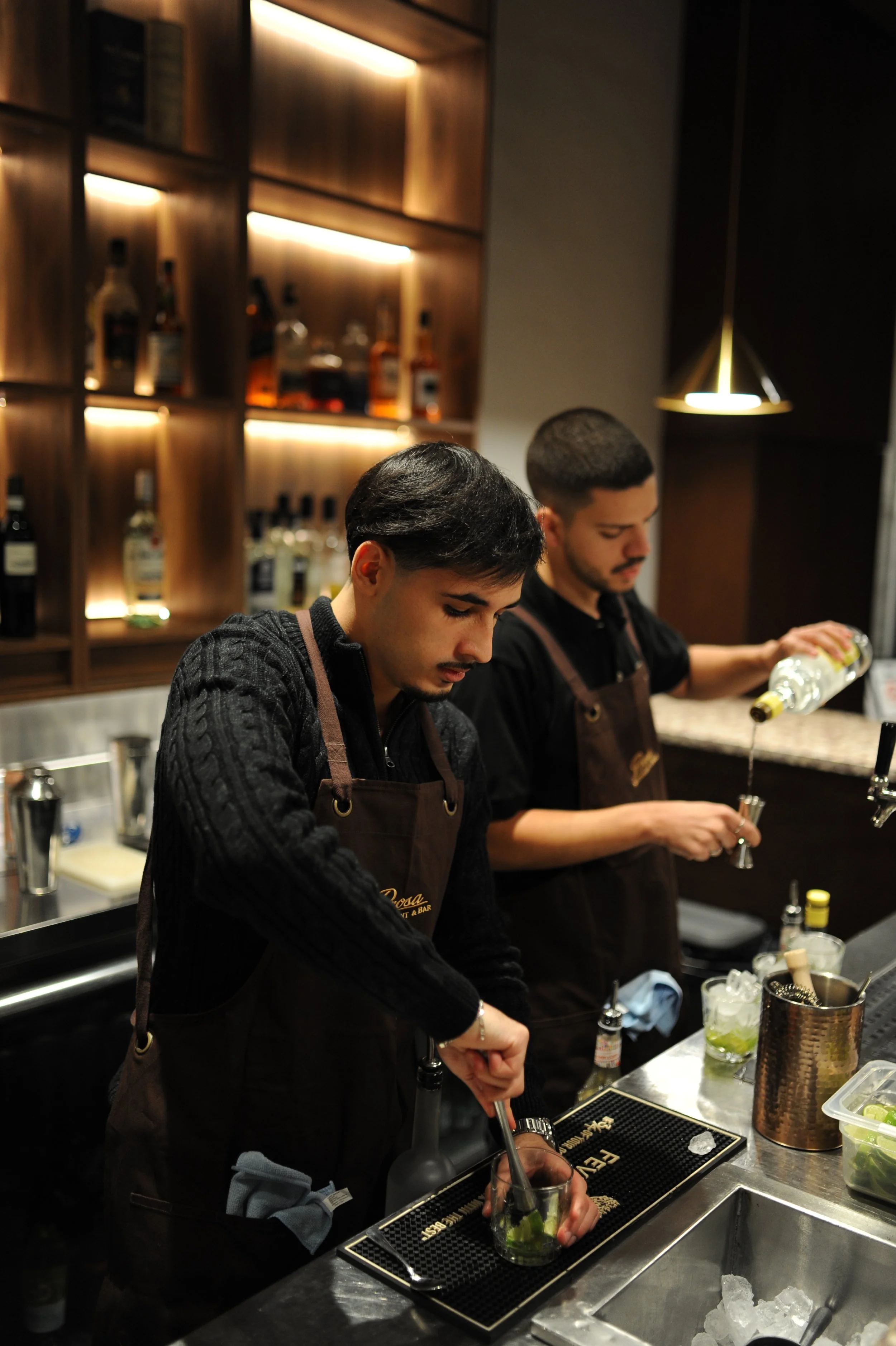 Two bartenders preparing drinks behind a bar, with shelves of liquor bottles illuminated in the background. One bartender is pouring a drink into a glass with ice and lime, while the other stirs a drink in a shaker.