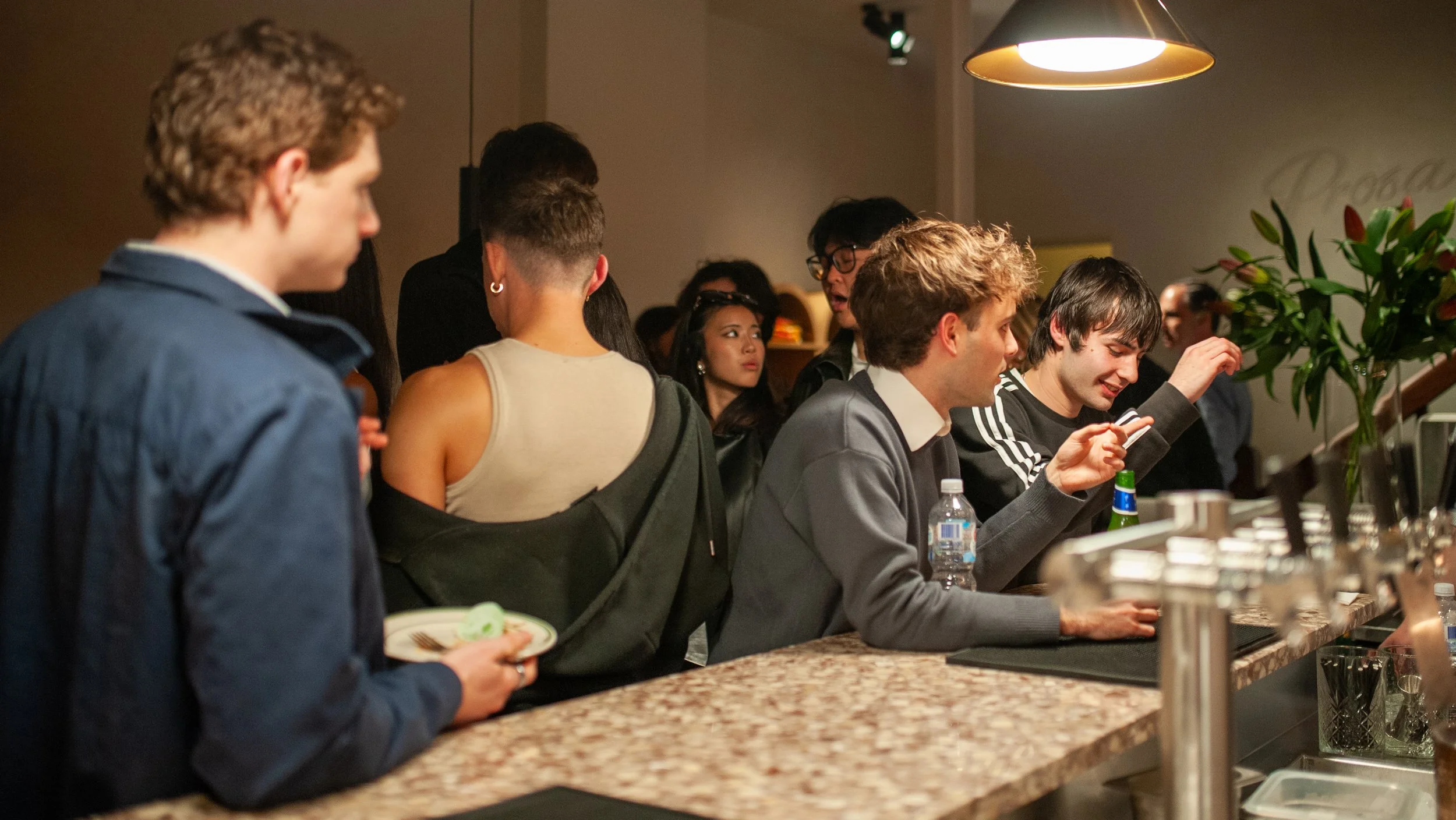 People standing at a bar or counter in a social setting, some looking at their phones, with a large plant and pendant lights in the background.