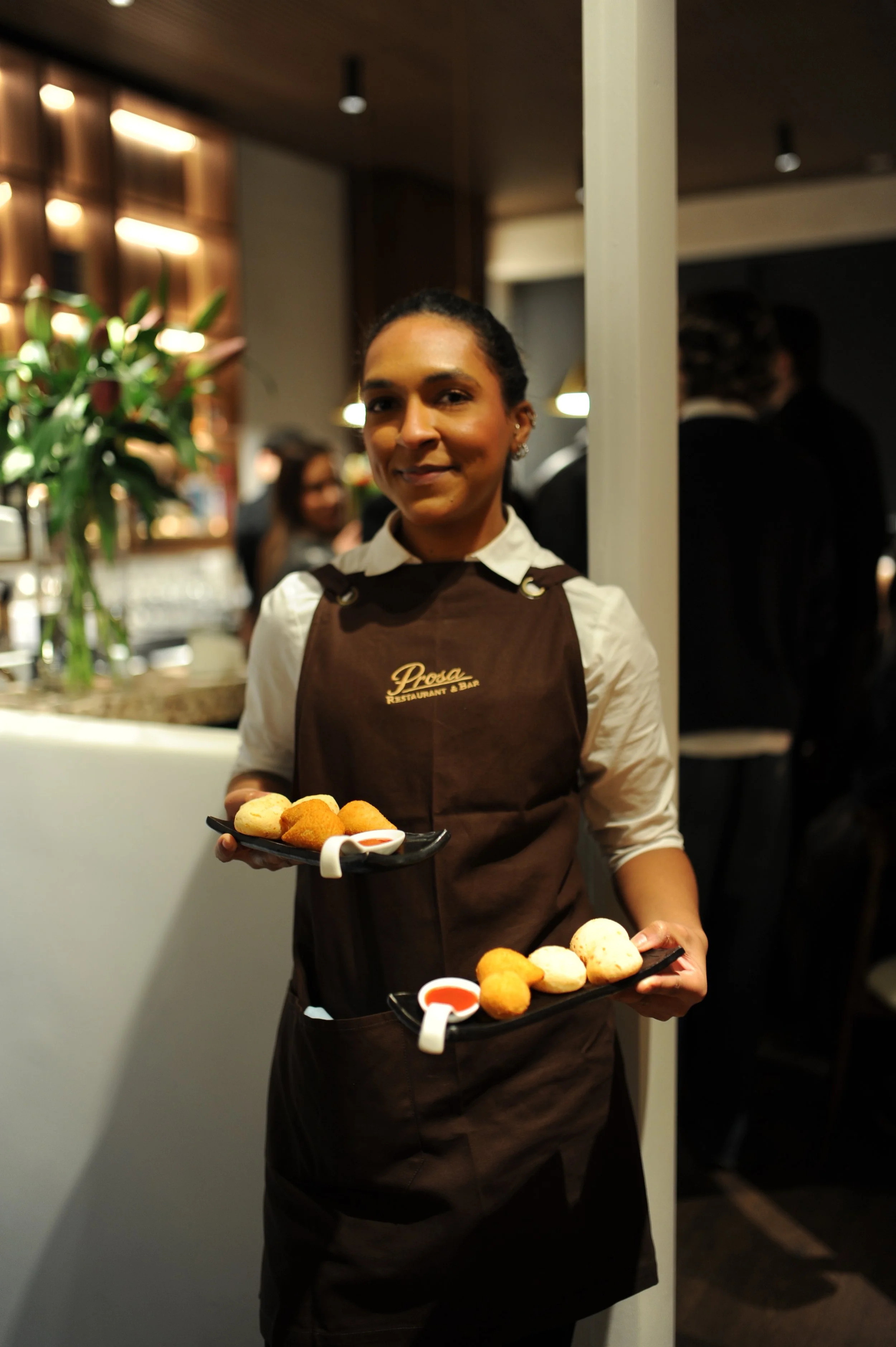 A smiling woman in a waitress uniform holding two trays of assorted fried appetizers, with a restaurant interior in the background.
