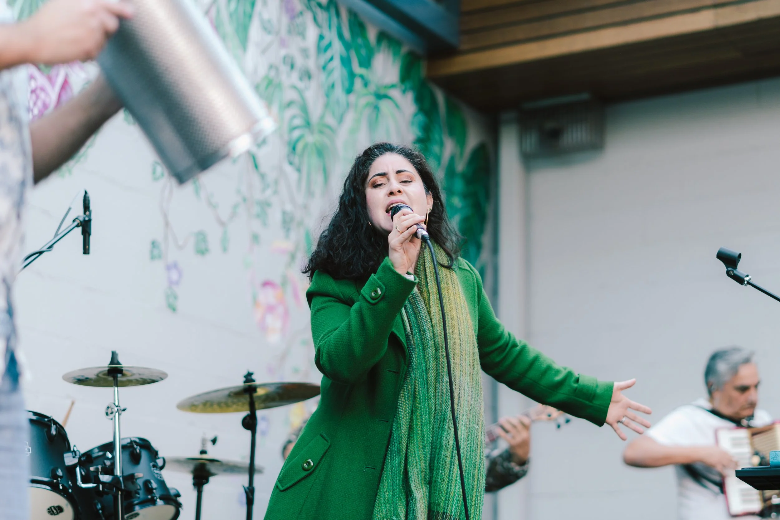 A woman singing into a microphone during a live outdoor music performance with musical instruments and other band members in the background.
