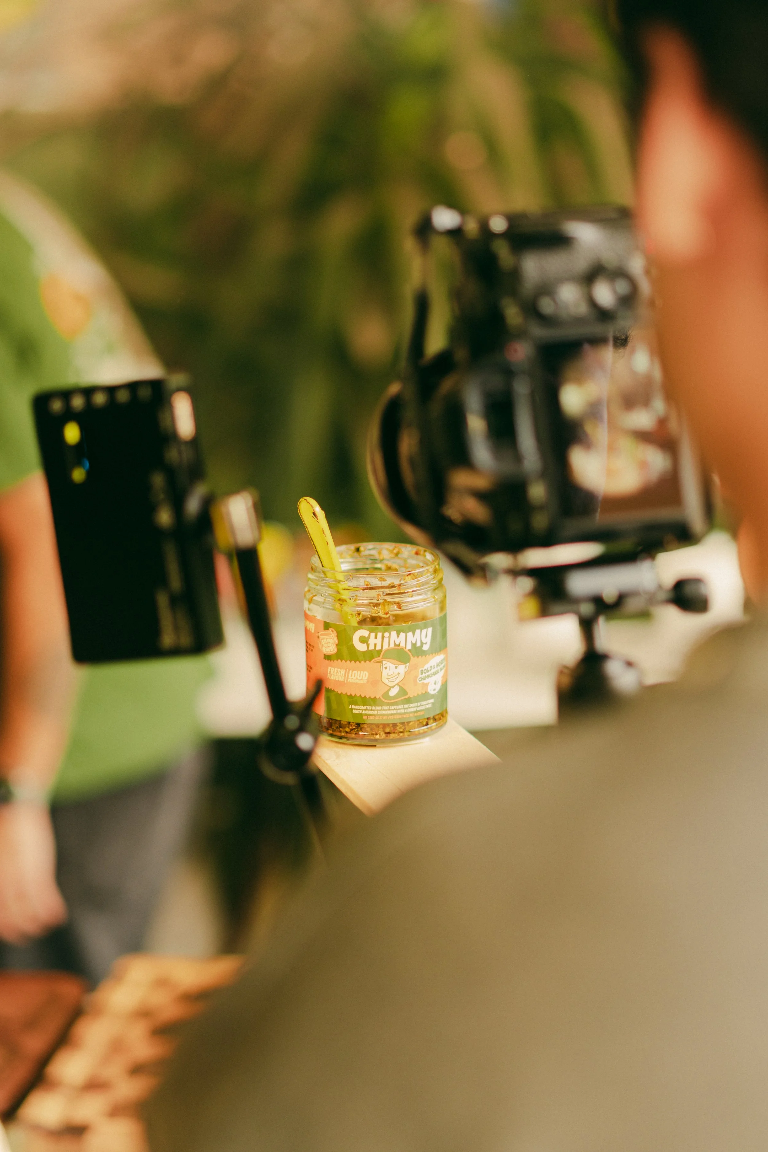 A camera is set up on a tripod focused on a jar of sauce labeled 'Chimmy' on a table.