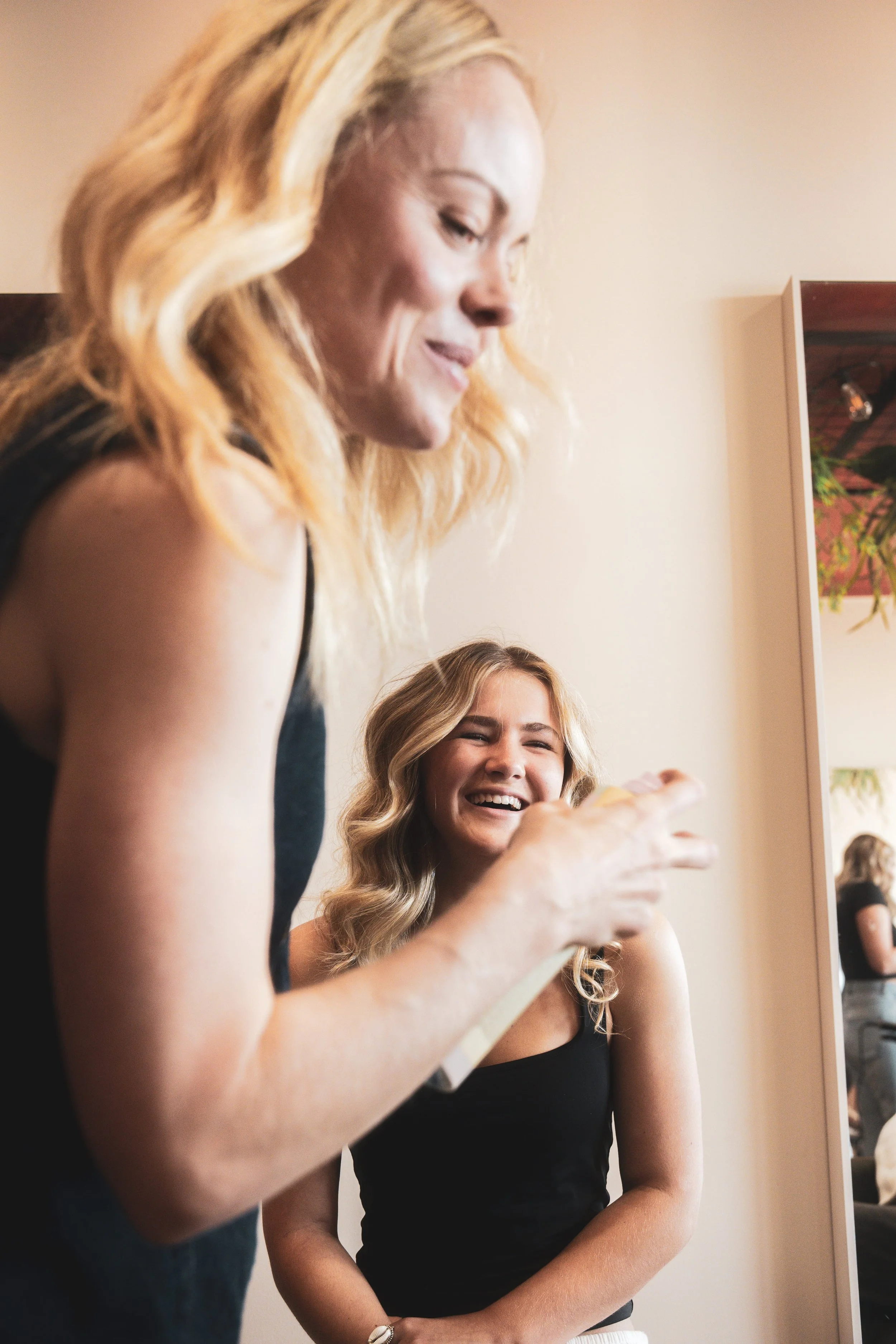 A woman with long blonde, wavy hair looking down and smiling, dressed in a black sleeveless top, standing next to another woman with light brown hair, also smiling, in a room with a beige wall and a wall-mounted mirror.