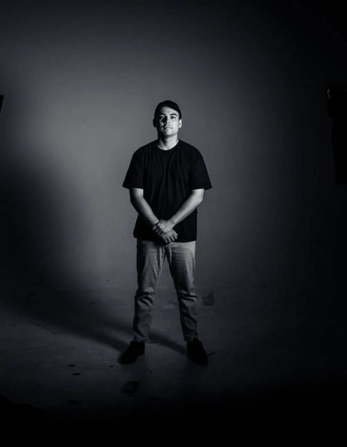 Black and white photo of a young man standing in a studio with a plain background, wearing a black t-shirt and light-colored jeans.