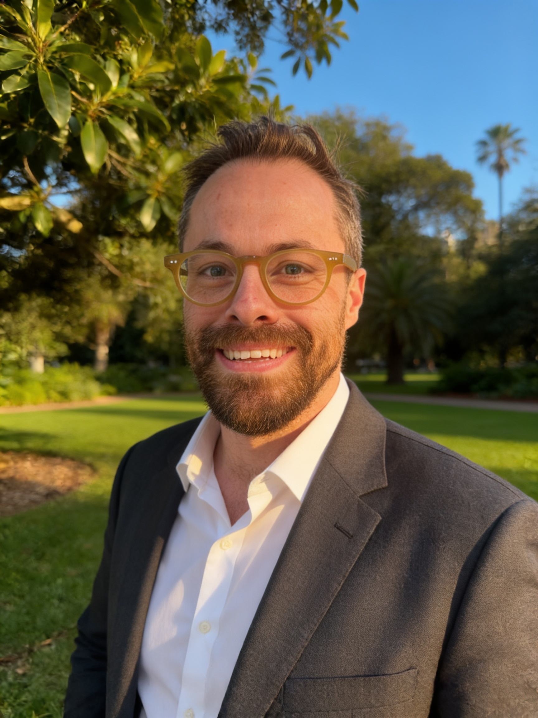 A smiling man wearing glasses, a white shirt, and a dark suit jacket, outdoors in a park with trees and a clear blue sky in the background.