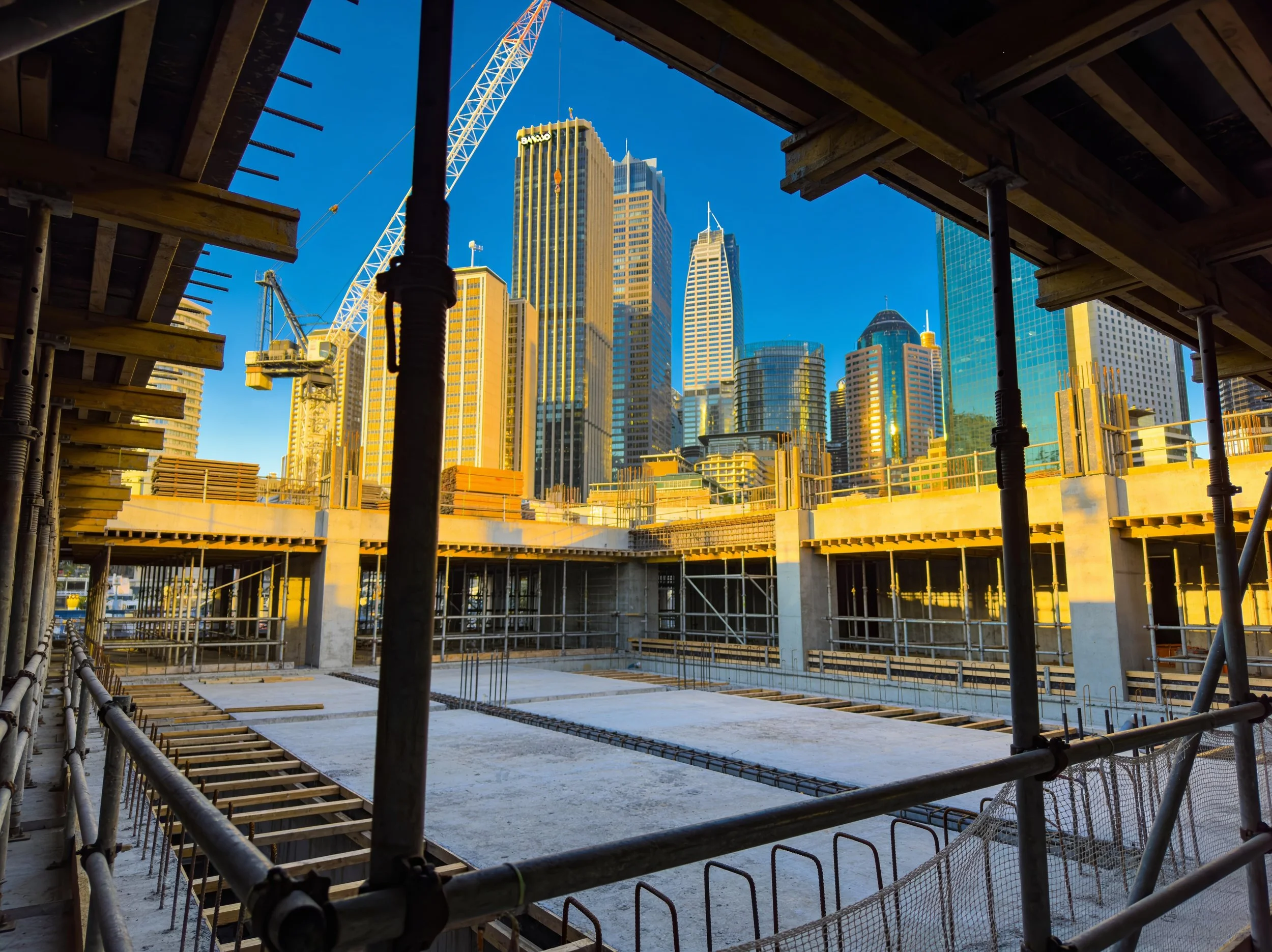 Construction site with scaffolding and concrete foundation, overlooking a city skyline with high-rise buildings and a blue sky.