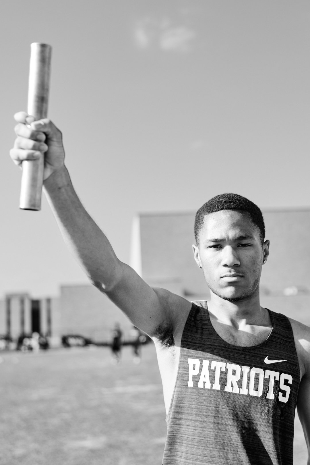 Black and white photo of a male athlete holding a baton in his right hand, standing on a sports field with a stadium in the background. He is wearing a sleeveless sports jersey with the word 'PATRIOTS' on it, looking serious.