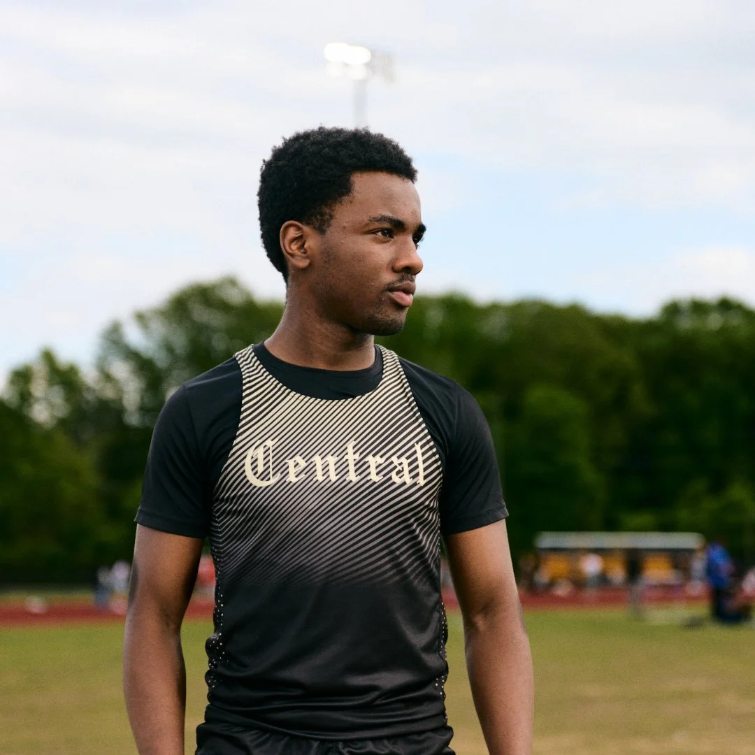 A young man with dark curly hair standing outdoors on a sports field wearing a black athletic shirt with "Central" written on it.