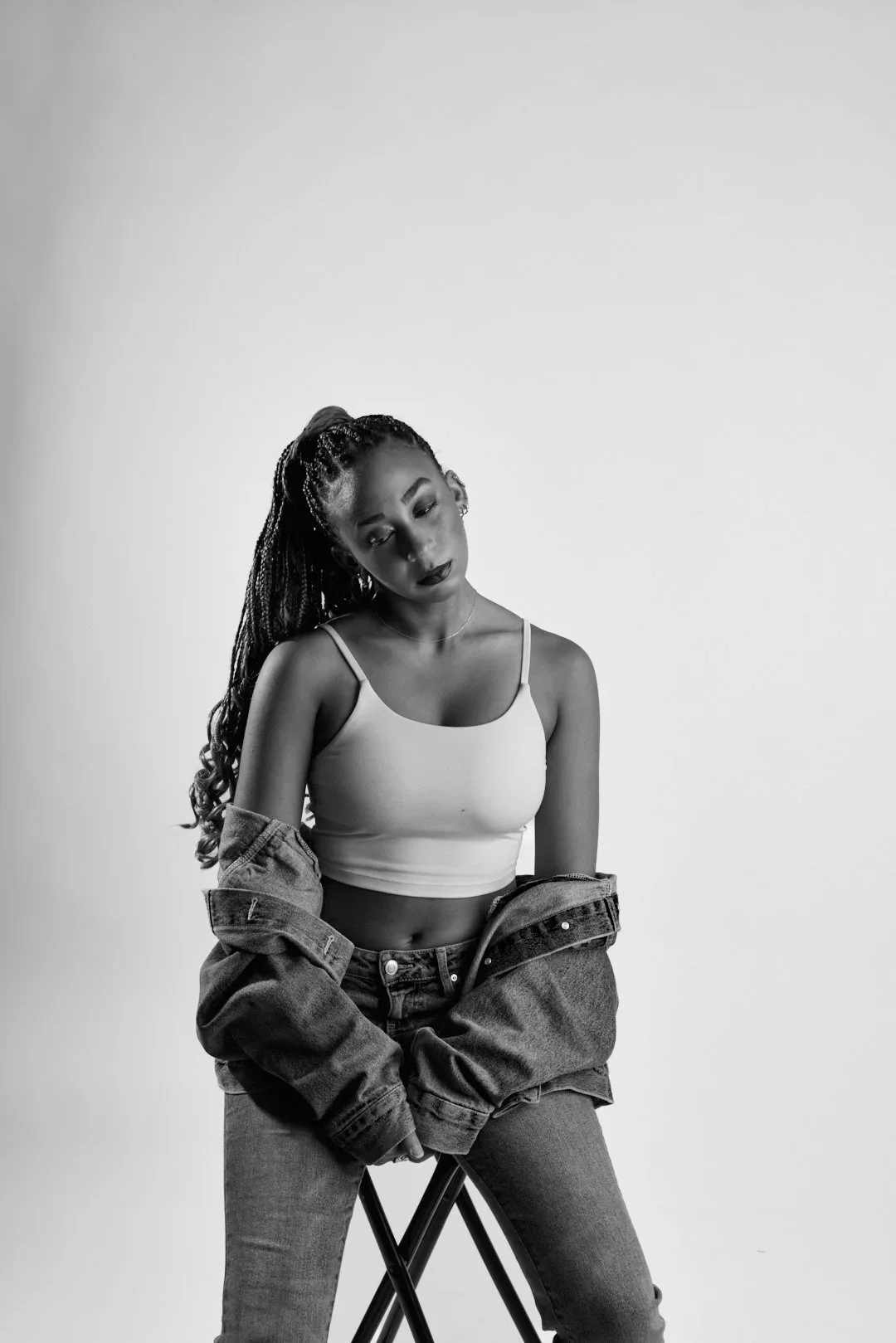 Black and white portrait of a young woman with long braided hair sitting on a stool. She wears a white tank top, jeans, and a jacket draped over her shoulders. She appears to be deep in thought with her head tilted downward.