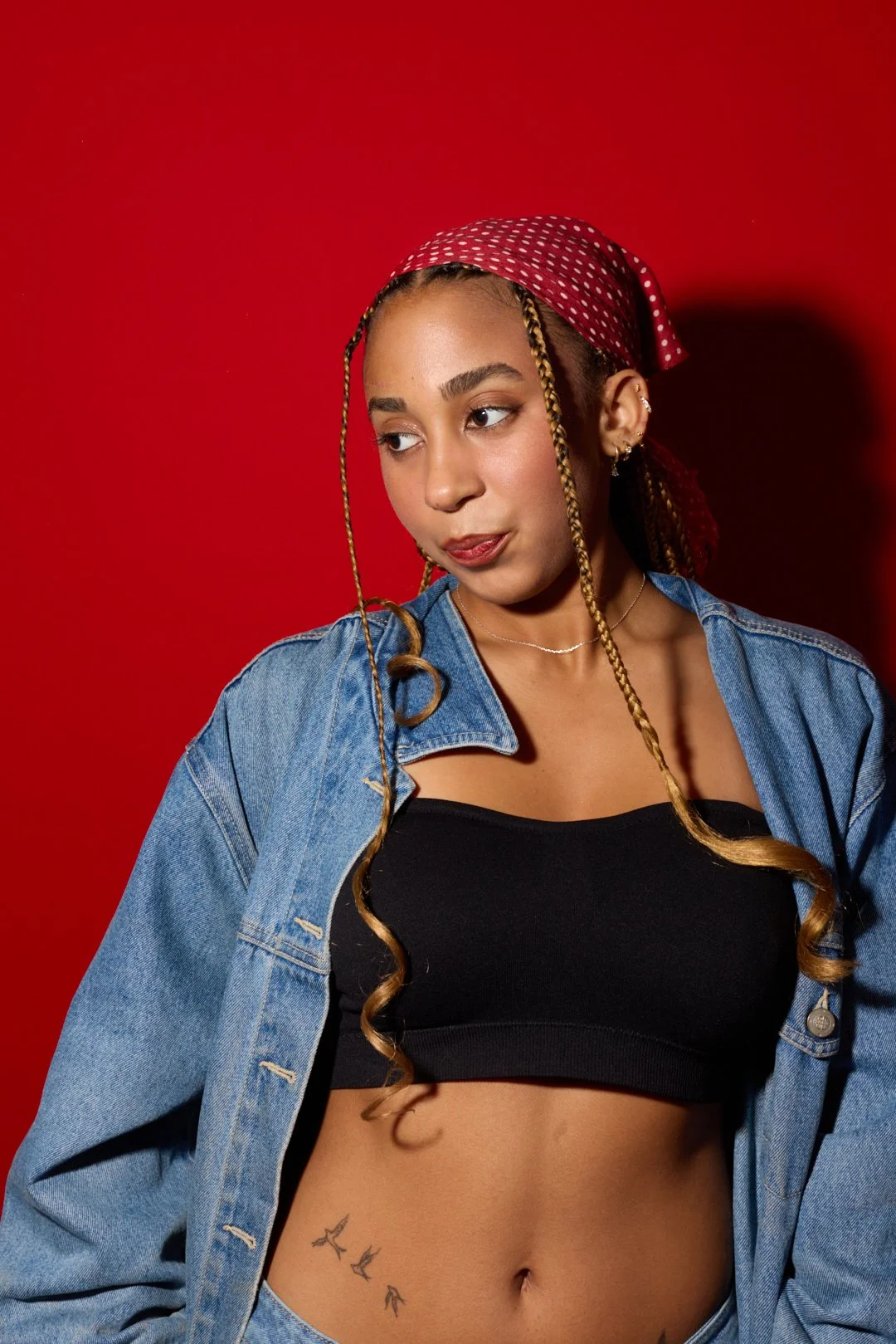 A young woman with long, braided hair, wearing a red polka dot headscarf, black crop top, and denim jacket against a red background.