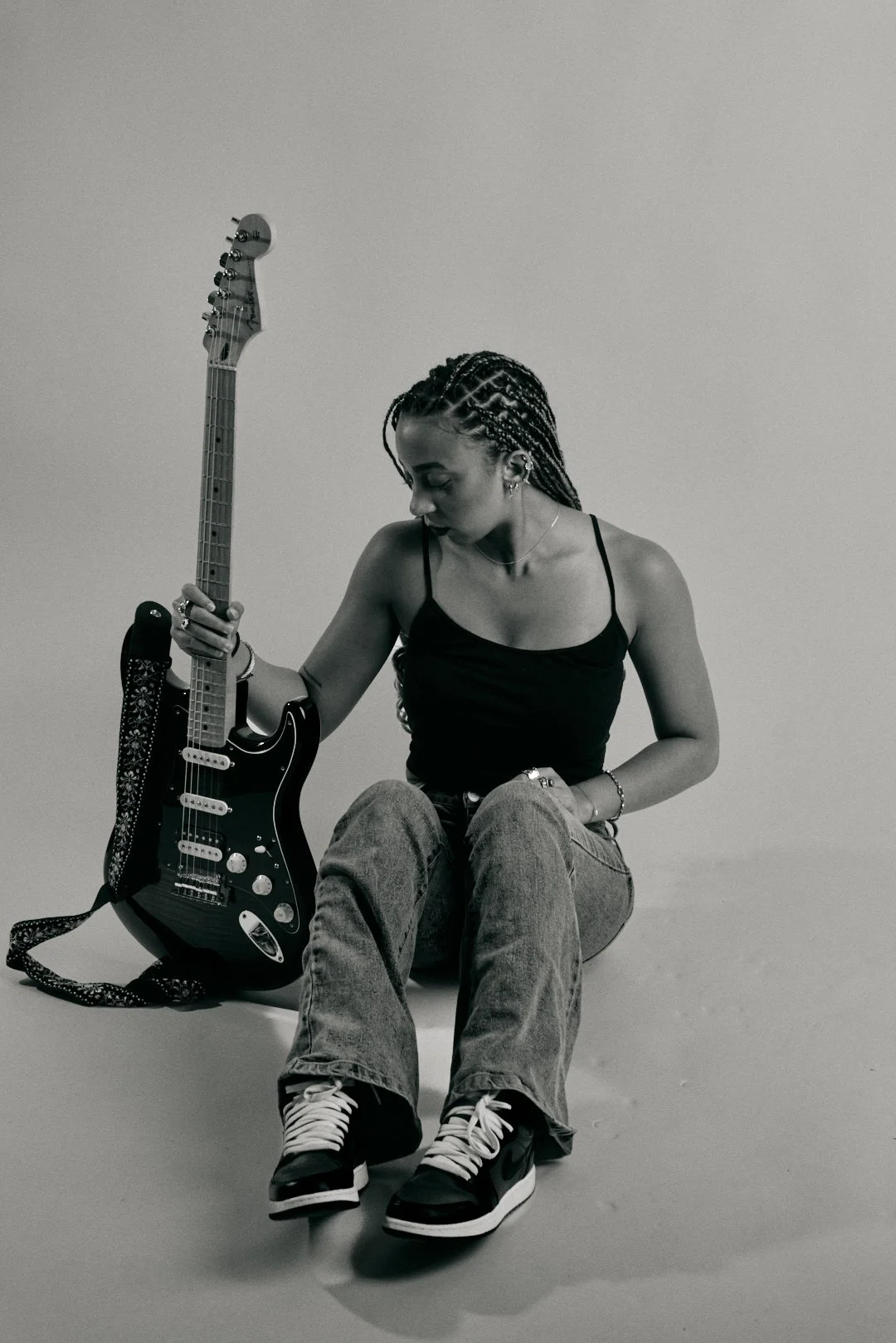 A woman with braided hair sitting on a white surface, holding an electric guitar, wearing a black tank top and gray jeans.