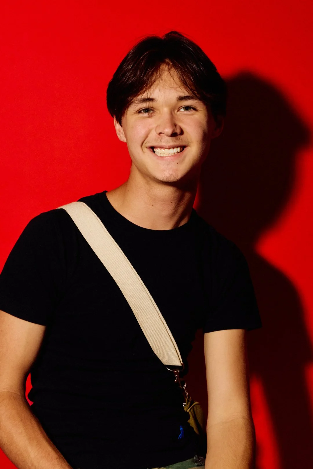 A young man with brown hair and a big smile, wearing a black t-shirt and a beige shoulder bag, standing against a red background.