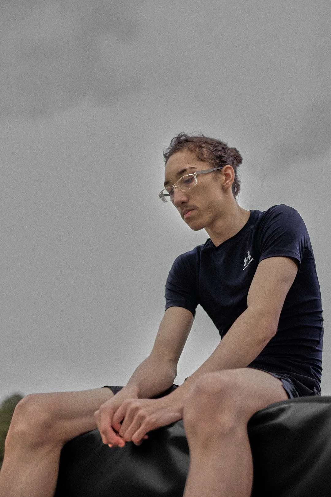 A person with glasses is sitting outdoors on a large black object, wearing a black t-shirt and shorts with an overcast sky in the background.