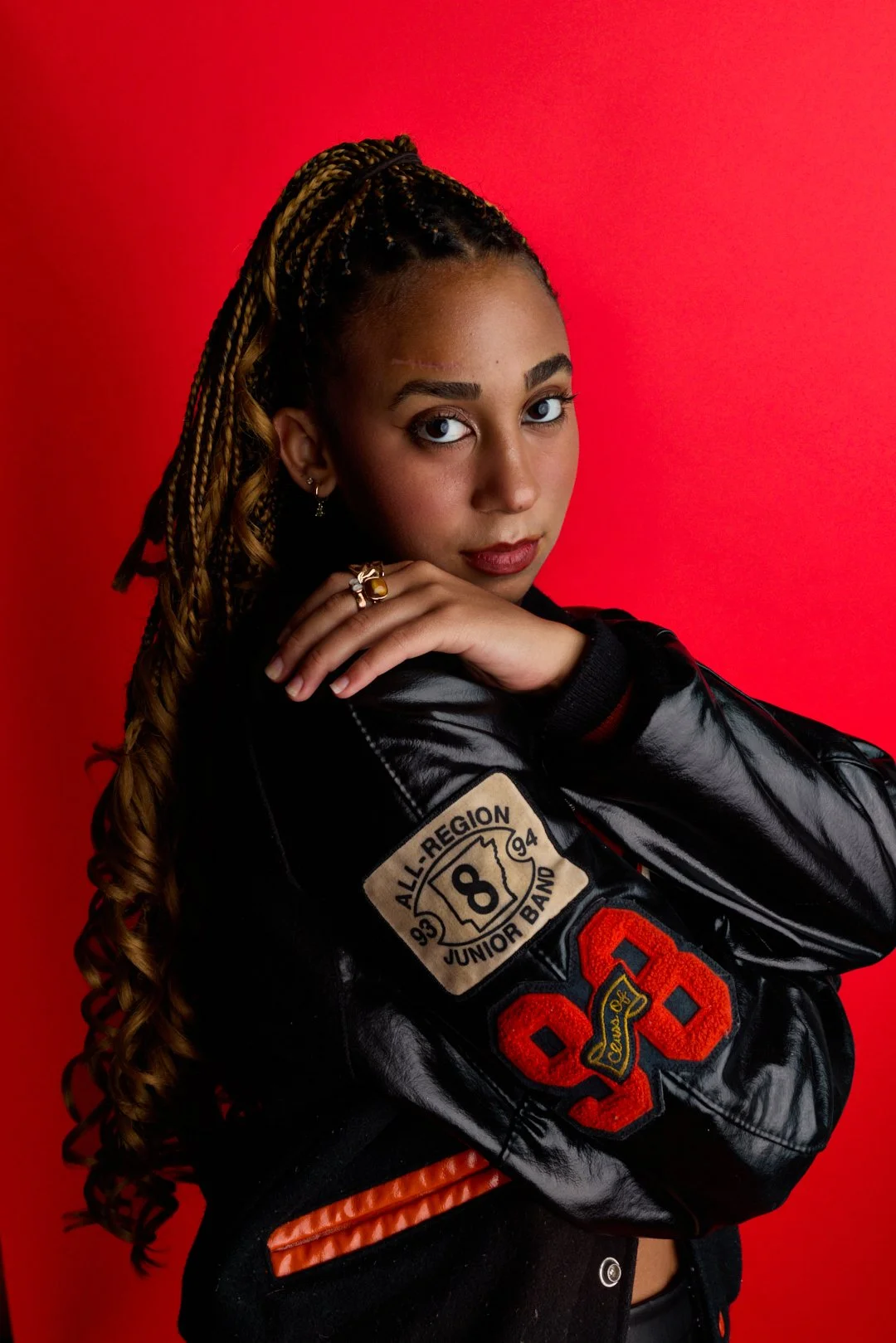 A young woman with light brown braided hair and light makeup, wearing a black shiny varsity jacket with patches, posing against a red background.