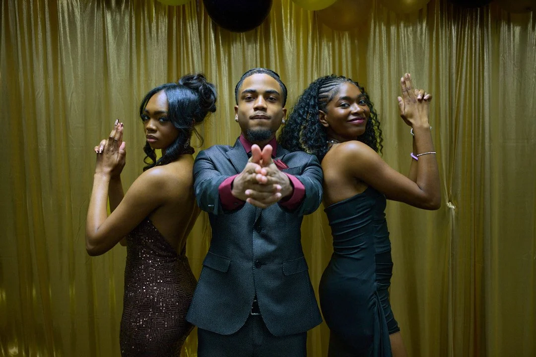 Three people posing back to back, dressed in formal attire, with a gold curtain background.