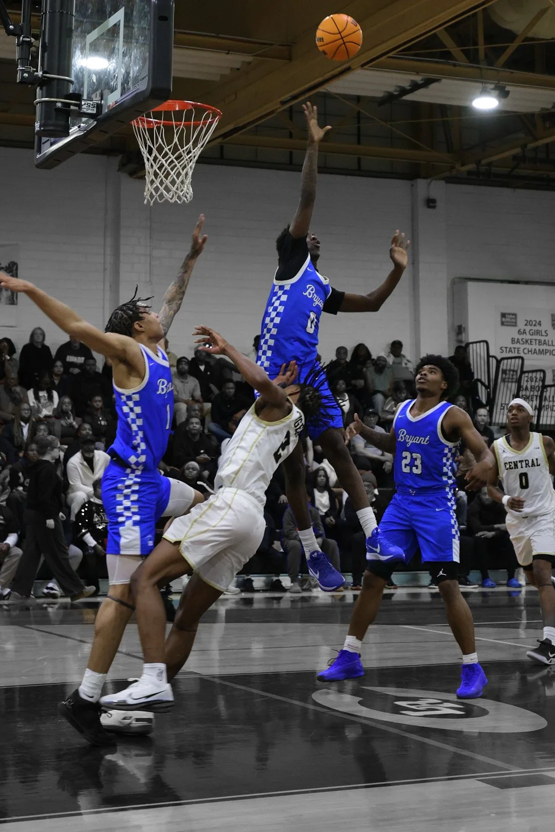 A men's basketball game in progress with players jumping for the ball near the basket. One player in a blue jersey is attempting a shot or block, while other players in blue and white jerseys surround him. The game is taking place in a gymnasium fill