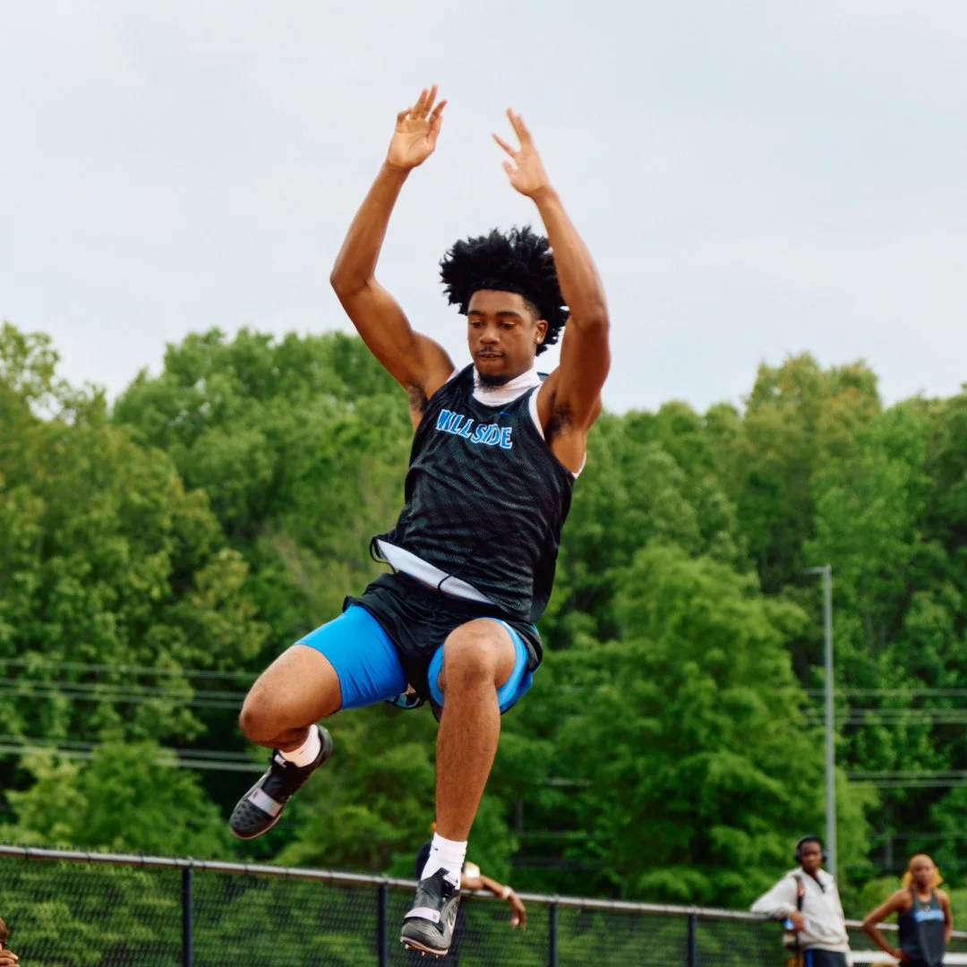 A young male athlete is jumping in the air during a track and field event, wearing a black and blue uniform with 'WILL'SIDE' written on it, with green trees in the background.