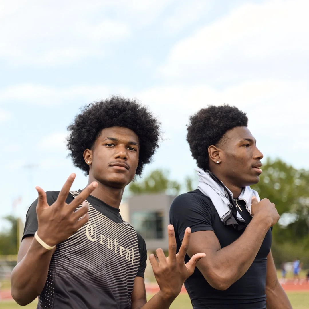 Two young men with athletic build and dark curly hair standing outdoors, one making a peace sign with his fingers and the other with a towel around his neck, on a track field during daytime.