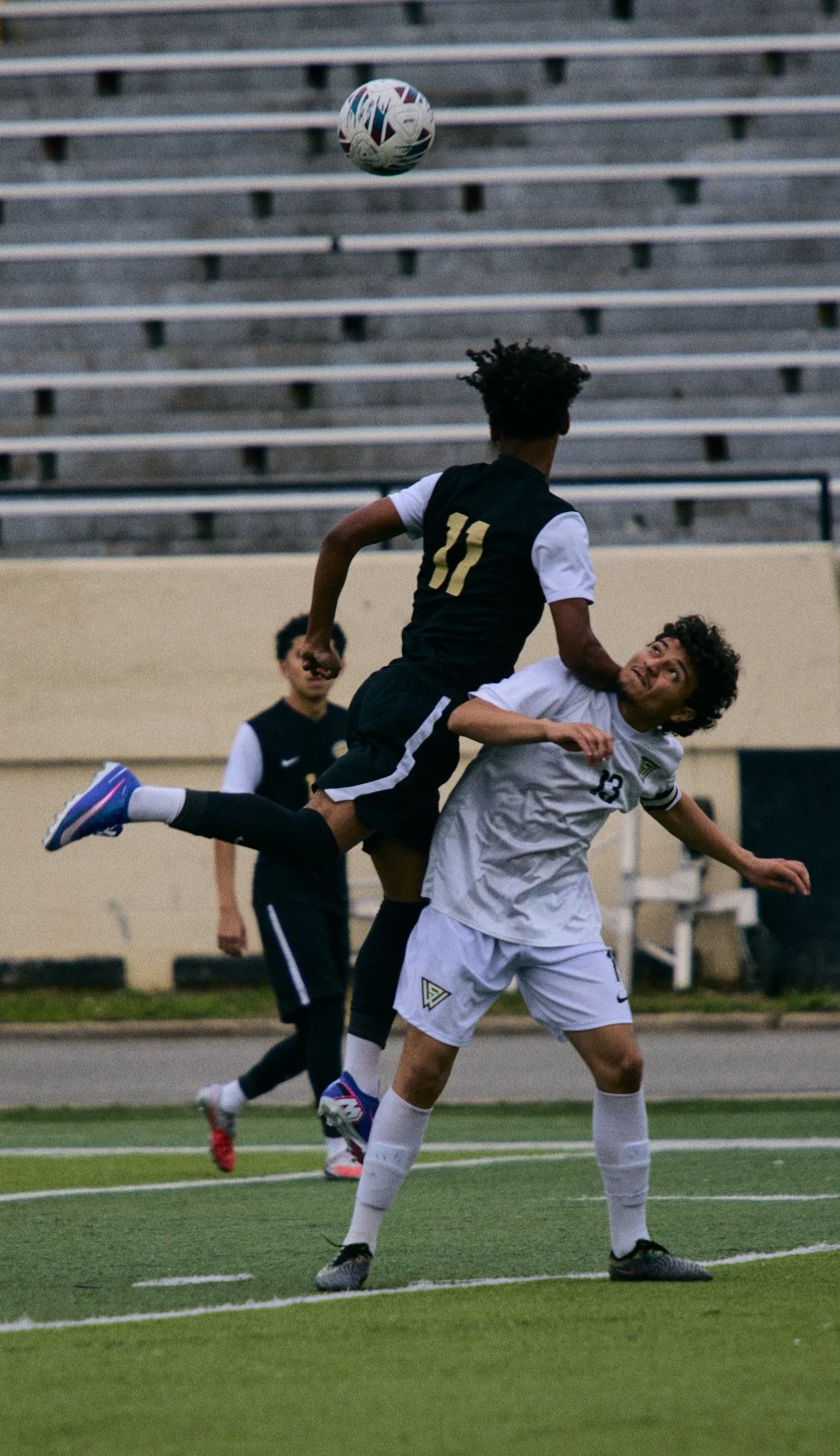 Two soccer players are competing for the ball on the field, with one player in a black jersey and the other in a white jersey. The black jersey player is jumping, and the white jersey player is attempting to block or challenge him. There is a third p