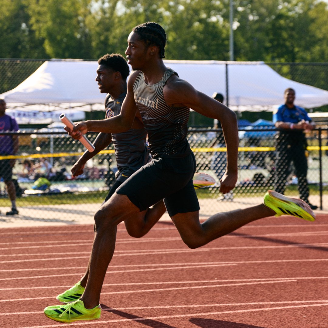 Two male runners competing on an outdoor track during daytime, with spectators and trees in the background.