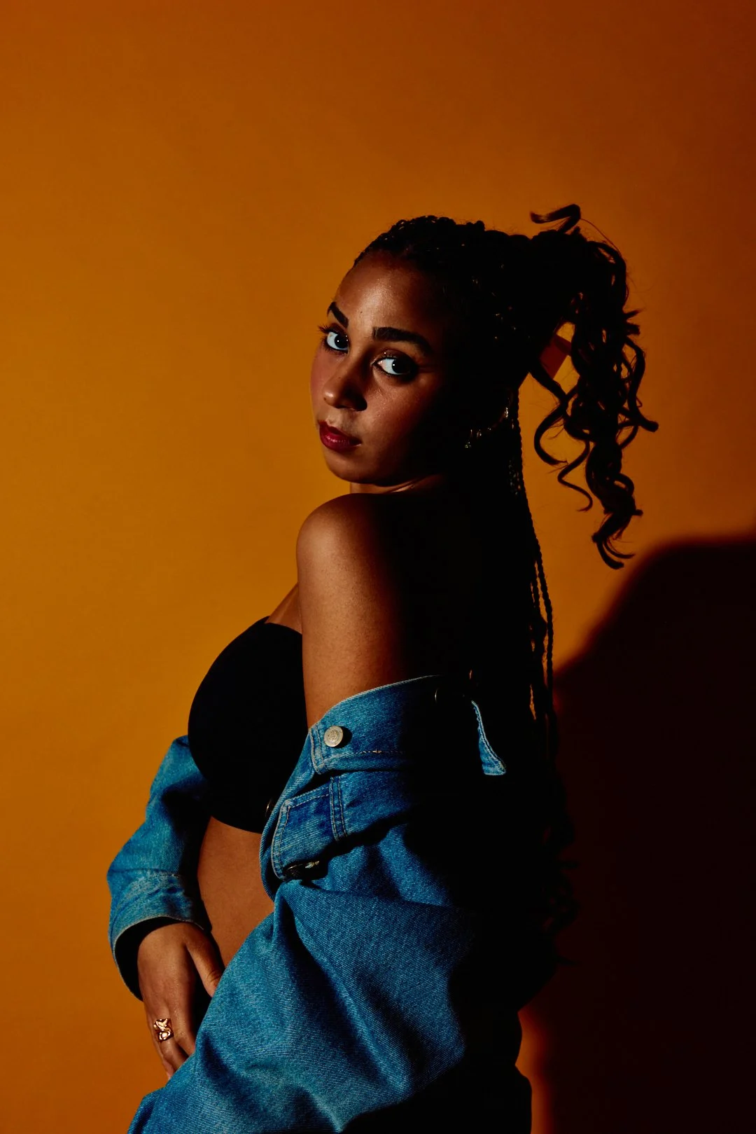 A young woman with dark, curly hair styled in a high ponytail, wearing a black strapless top and a blue denim jacket that is slipping off her shoulders. She is standing against a warm orange background, with her gaze directed towards the camera.