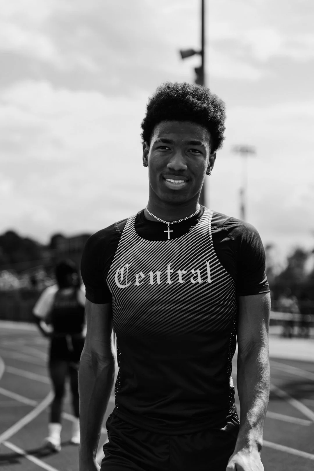 A young male athlete with curly hair and earrings, wearing a sports jersey with 'Central' written on it, standing on an outdoor track with a blurred background and a cloudy sky.