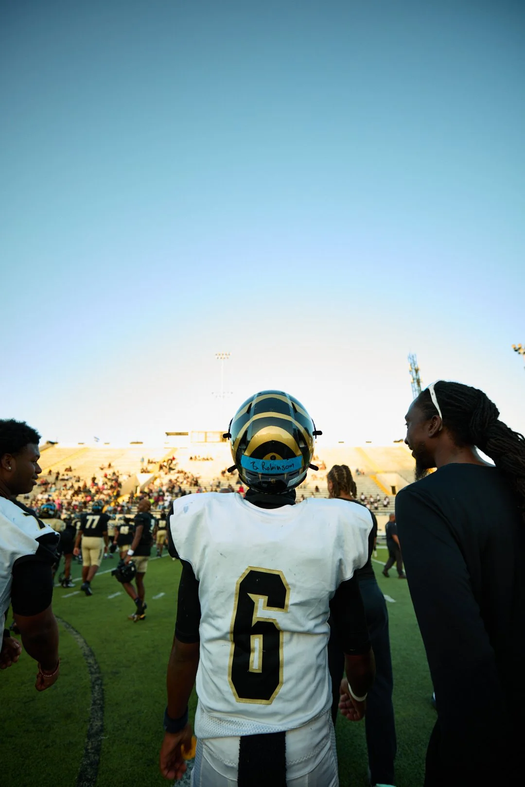 A football player wearing a helmet and white jersey with the number 6, standing on the field with other players and a man, during daytime at a stadium with a crowd in the background.