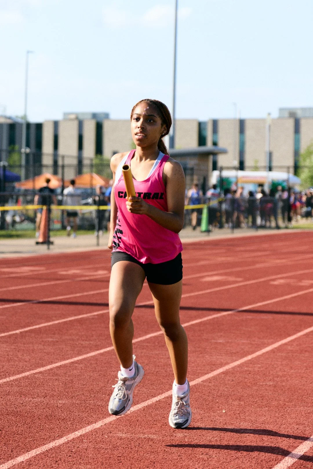 A young female athlete running on a red track during a relay race, holding a baton in her right hand, with a crowd and tents in the background.