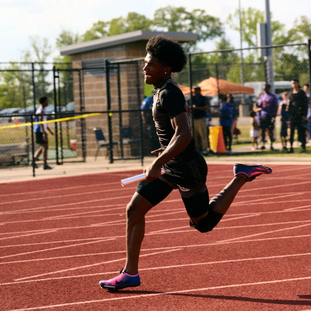 A young male athlete running on a red track at an outdoor sports field, holding a relay baton in his right hand, with spectators and field facilities in the background on a sunny day.