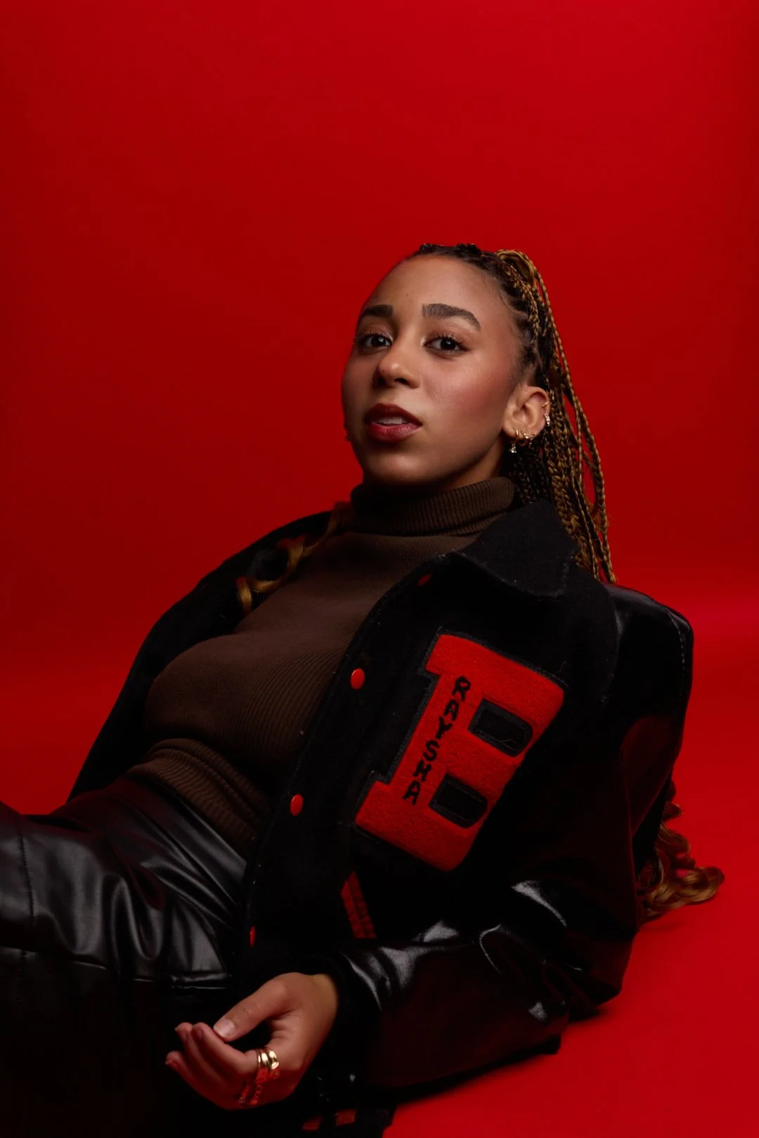 A young woman wearing a black jacket with letter patches and a brown turtleneck, posing against a red background.
