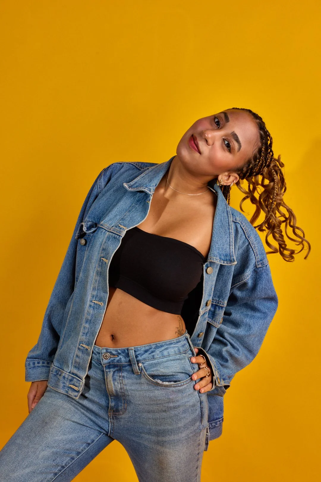 Young woman with curly hair styled in braids, wearing a denim jacket over a black crop top and jeans, standing against a yellow background.
