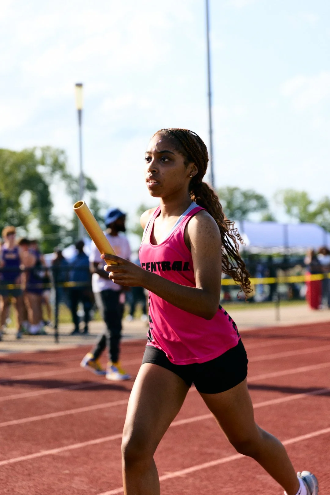Young female athlete in pink race uniform running on track during relay race holding baton at outdoor stadium with spectators in background.