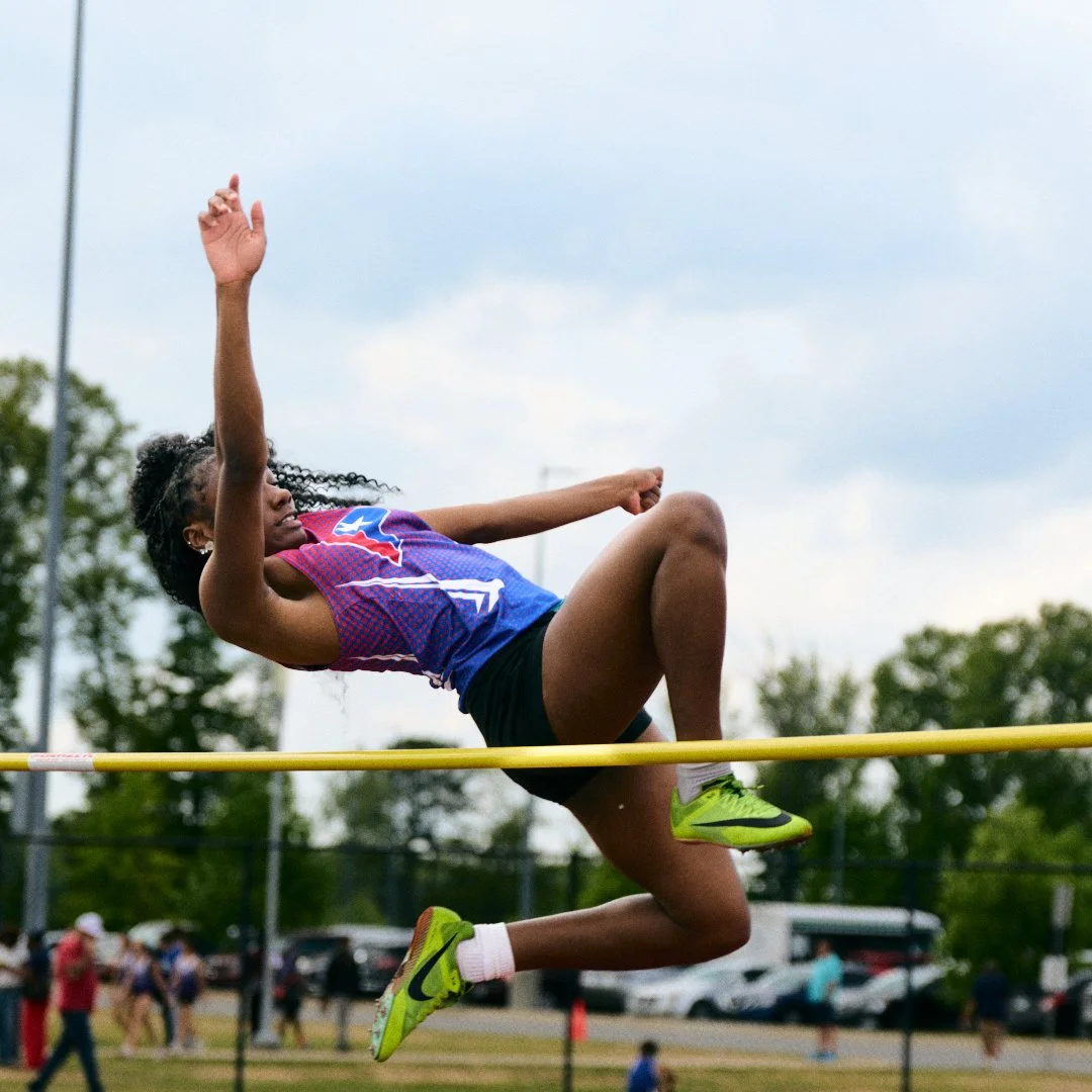 Girl in a sports uniform jumping over a high jump bar during a track and field event.