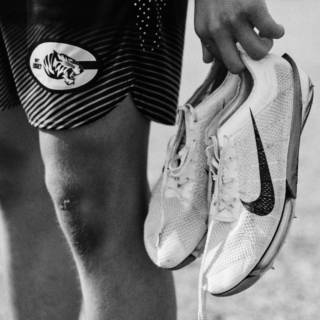 A person wearing striped athletic shorts displaying a logo, holding a pair of running shoes by the laces, with the focus on the shoes.
