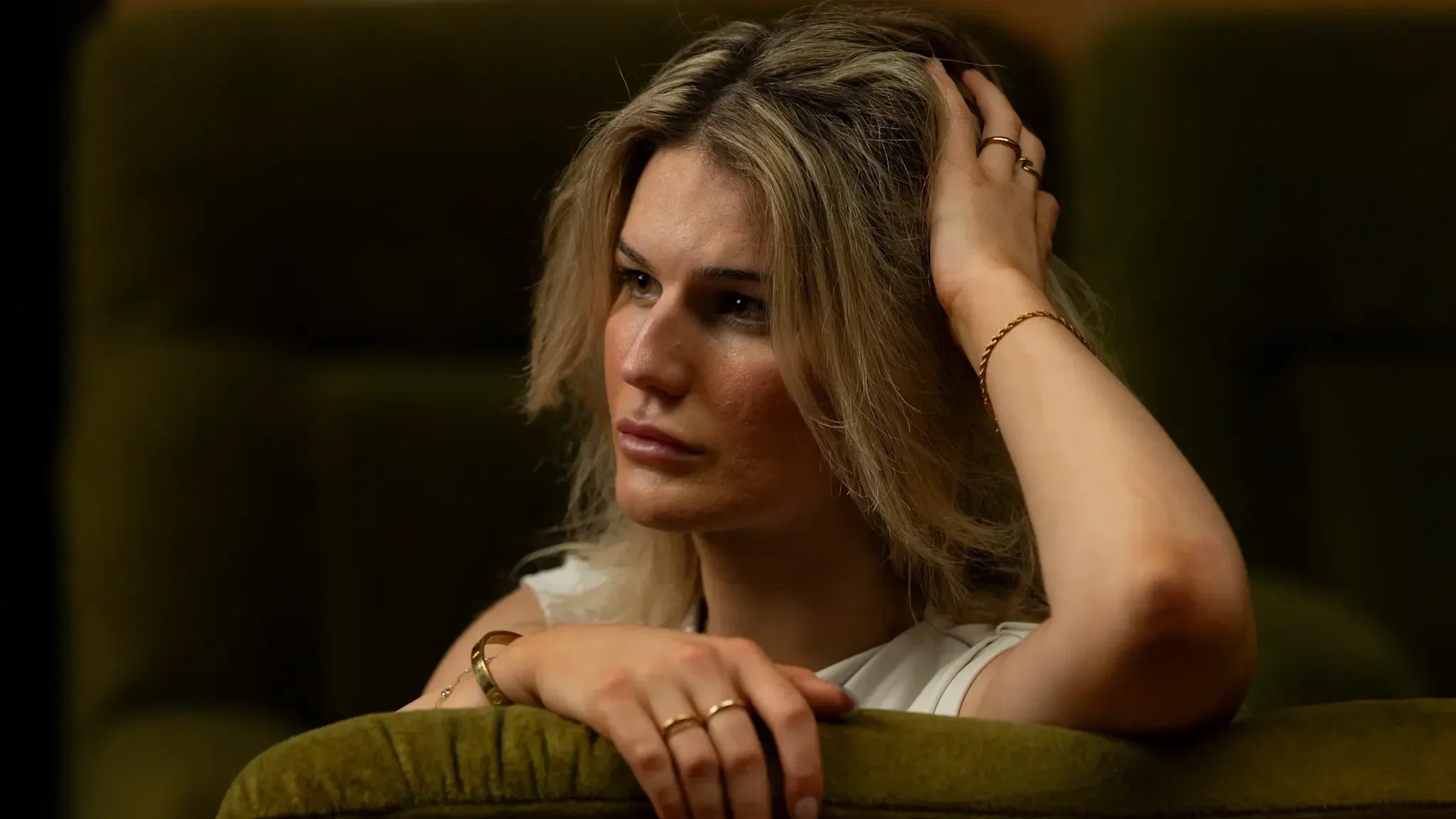A woman with blonde hair resting her hand on her head while sitting on a green velvet chair, with a serious expression on her face.