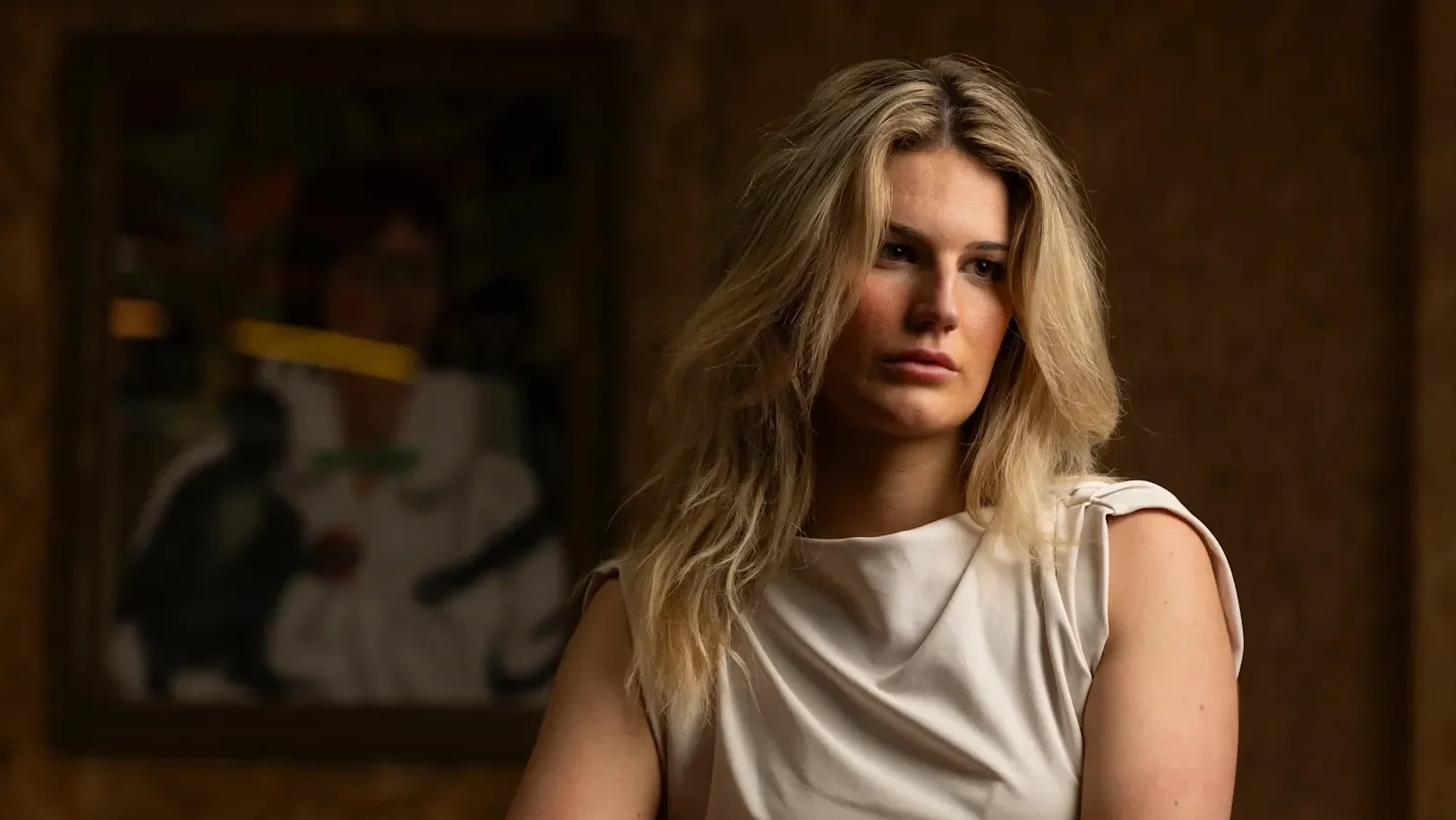 A woman with blonde hair and a white sleeveless top, looking contemplative, indoors with a blurred abstract painting in the background.