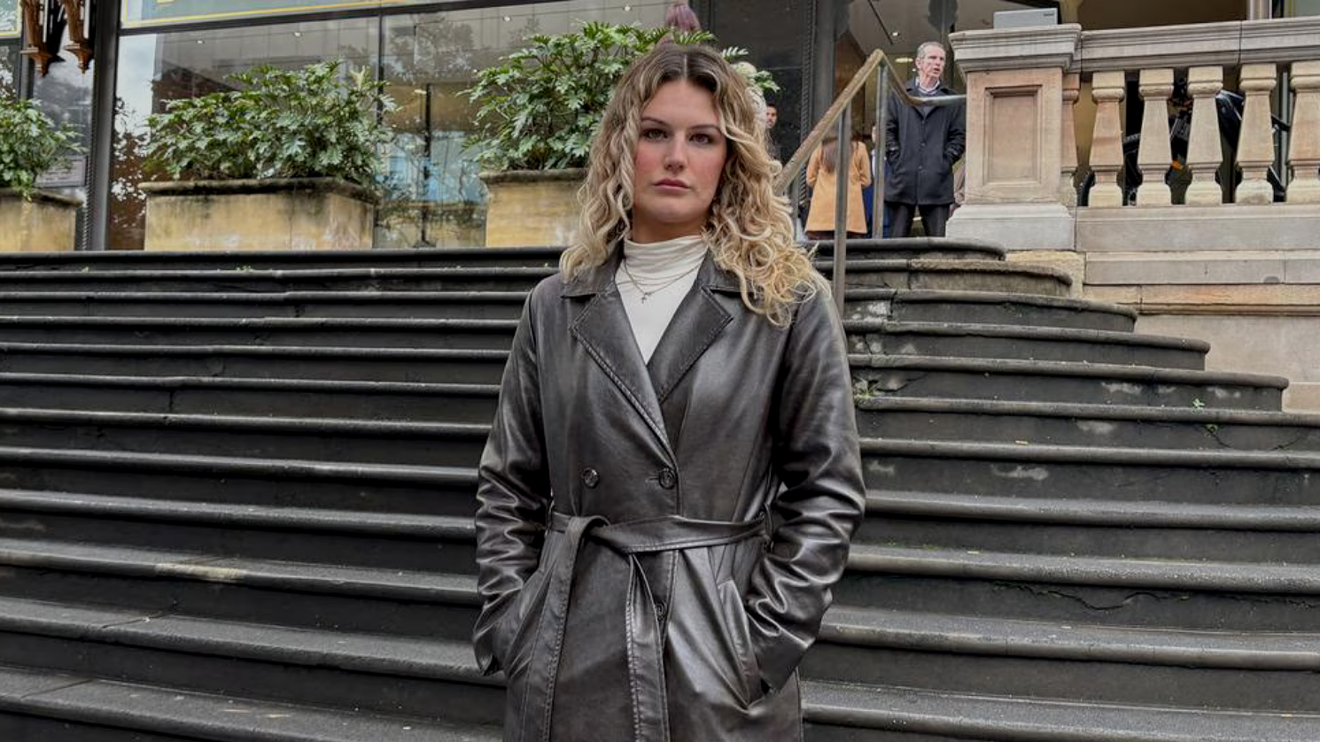 A young woman with curly blonde hair standing on city stairs wearing a black leather trench coat.