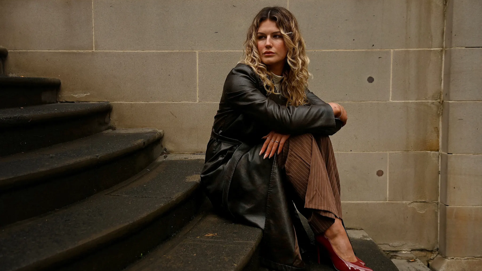 Woman with curly blonde hair sitting on stairs against a stone wall, wearing a black leather jacket, striped brown pants, and red high heels.