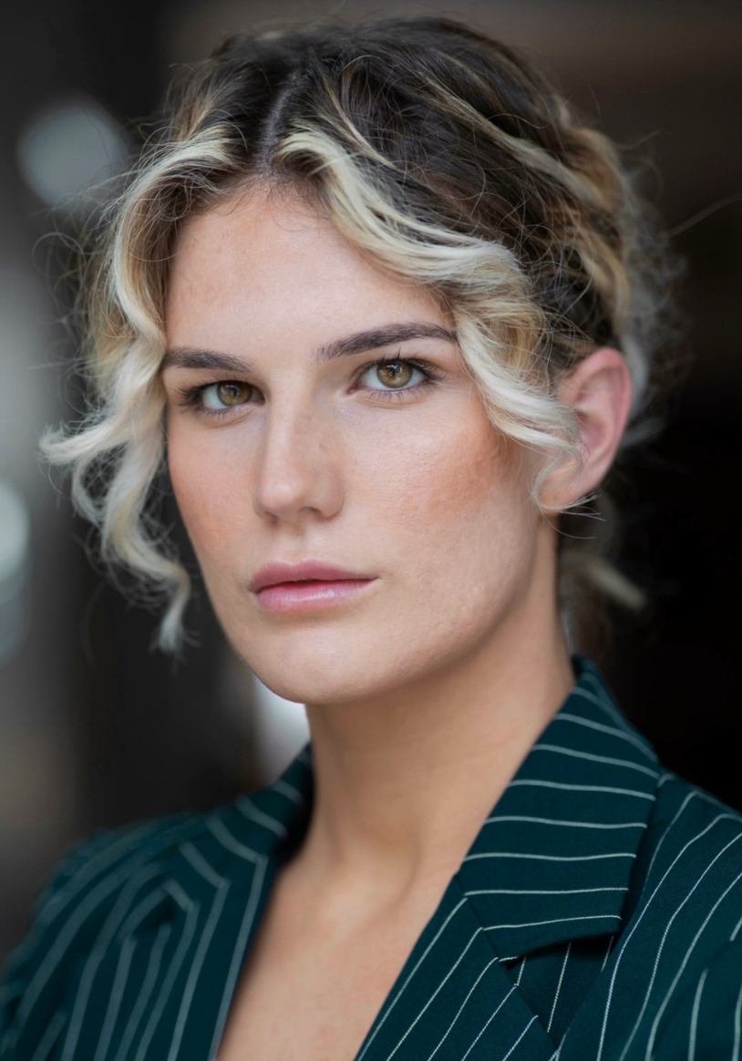 Close-up portrait of a woman with short, curly blonde hair and hazel eyes, wearing a dark pinstripe blazer.