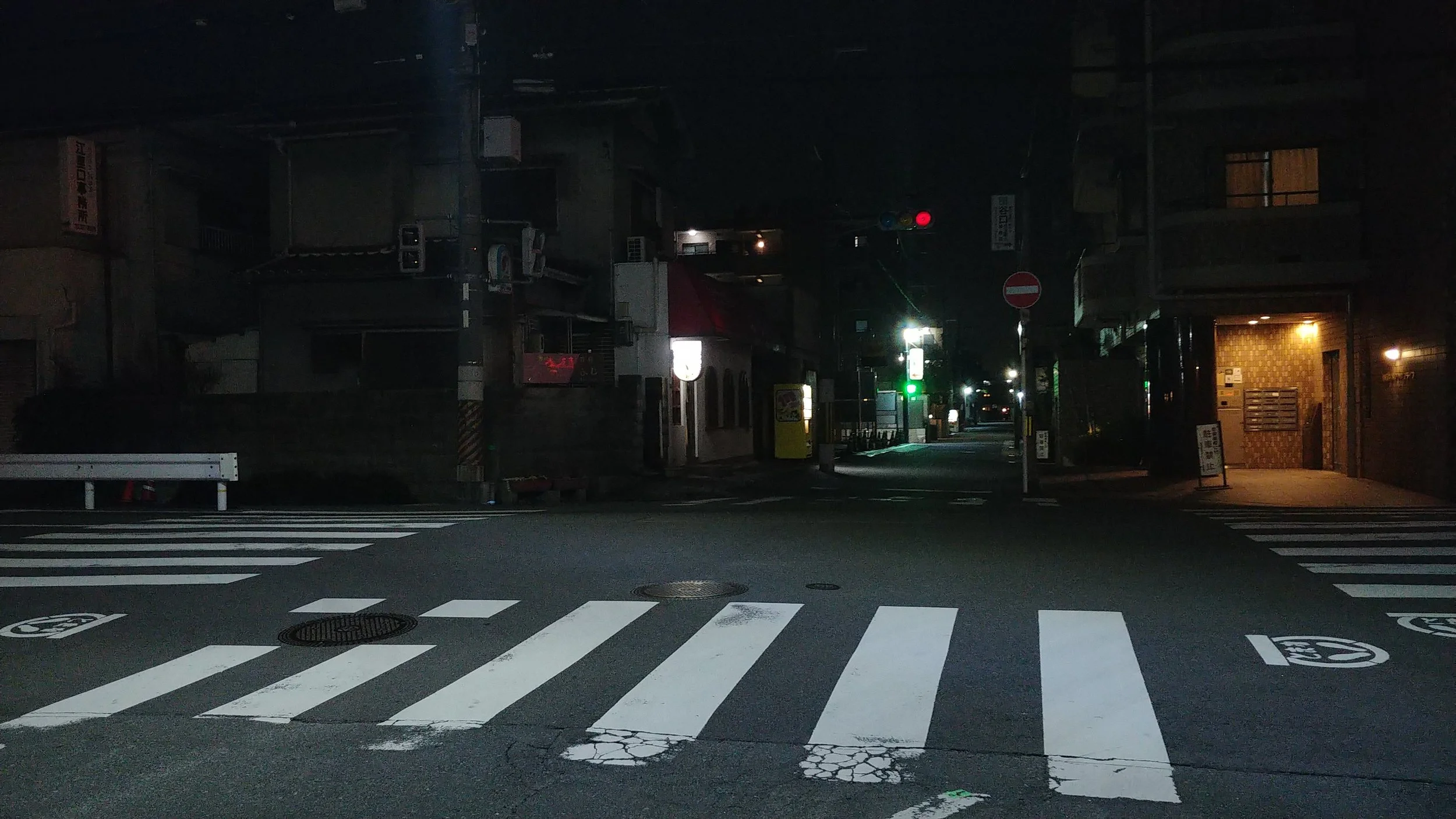 Nighttime urban street scene with a pedestrian crosswalk and traffic light. Commercial buildings are visible on the sides, some with lit windows and signs.