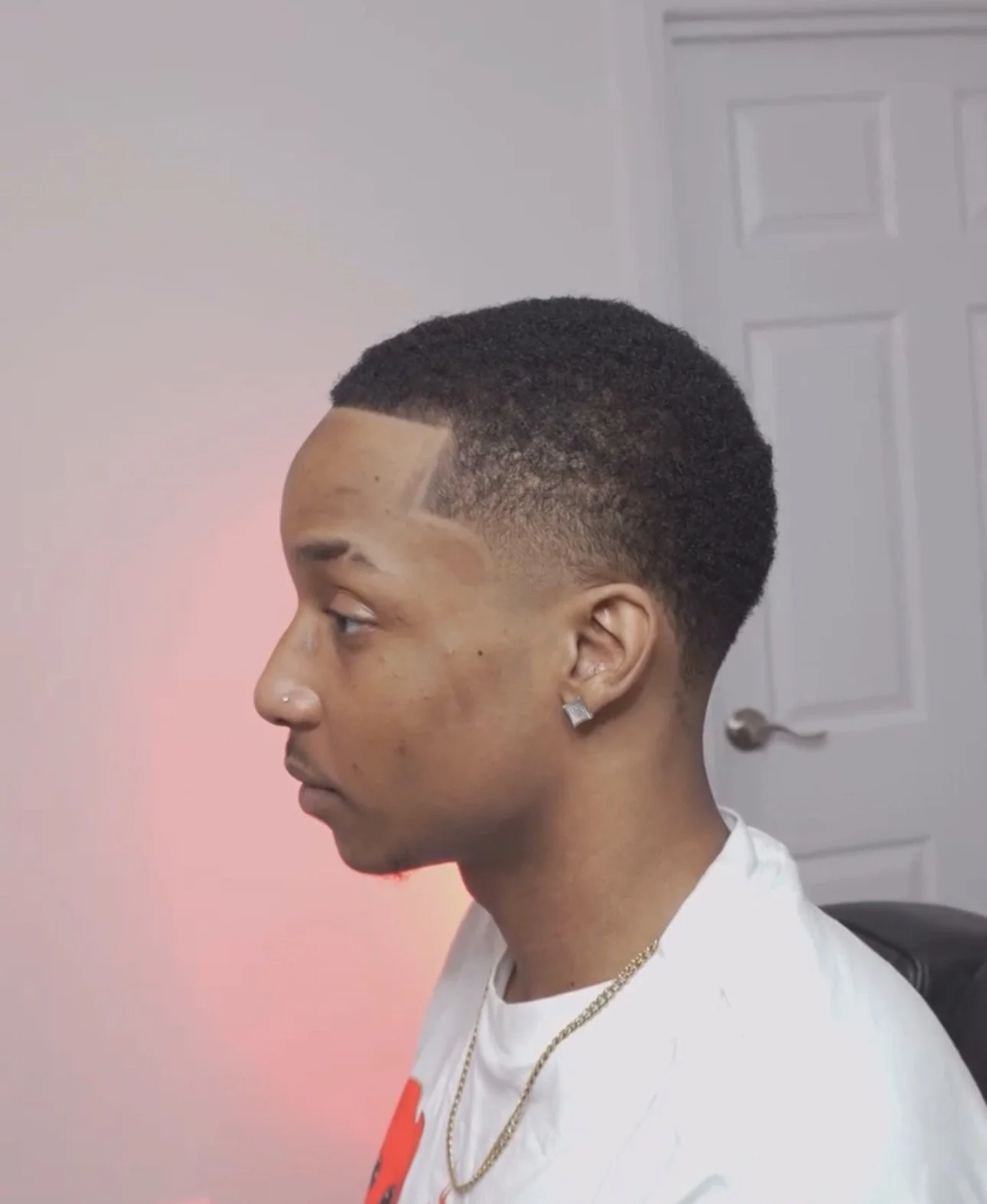 Profile of a young man with a fresh haircut featuring a geometric design shaved into the side of his head, wearing earrings, a nose piercing, a gold chain, and a white shirt, sitting in a room with a white door in the background.